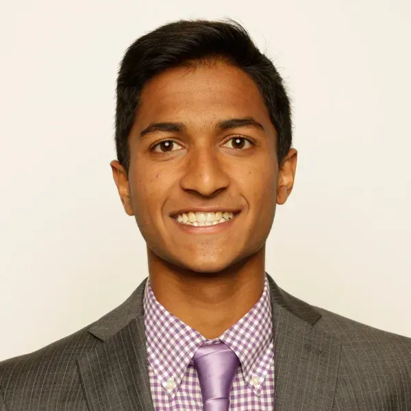 Smiling young man in a gray suit, purple checked shirt and lavender tie, against a plain background