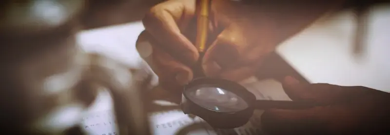 A person holding a pencil examines a printed page through a magnifying glass in warm, dim light.