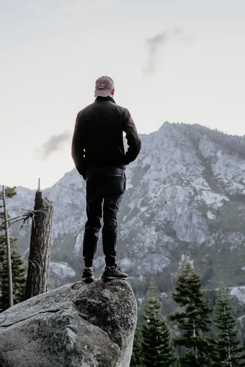 A person in a cap and dark jacket stands on a boulder, facing a snow-dusted mountain and pine trees.