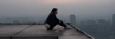 A young woman sits alone on a rooftop edge, looking out over a hazy city skyline at dusk.