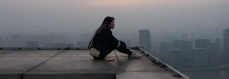 A young woman sits alone on a rooftop edge, looking out over a hazy city skyline at dusk.