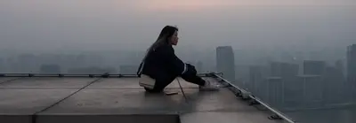 A young woman sits alone on a rooftop edge, looking out over a hazy city skyline at dusk.