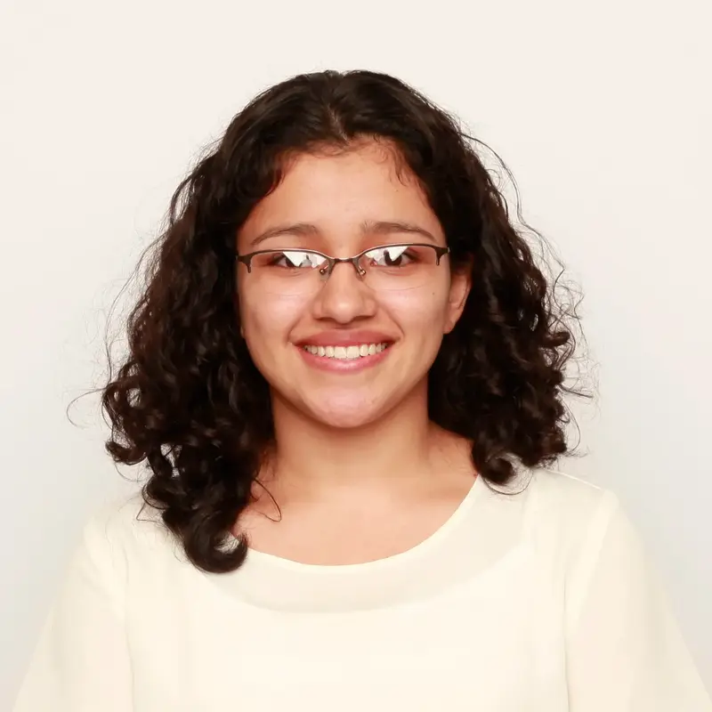 Smiling student with curly dark hair and glasses in a cream top against a light background