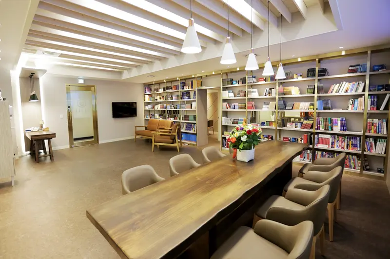 A modern study lounge with a long wooden table, upholstered chairs, pendant lights and floor-to-ceiling bookshelves