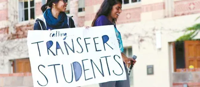 "calling... TRANSFER STUDENTS"라고 쓴 손글씨 팻말을 들고 미소 짓는 두 명의 학생