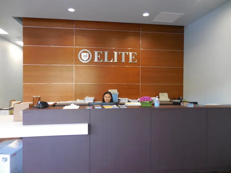 A smiling receptionist at a front desk beneath an "ELITE" sign on a wood-paneled wall.
