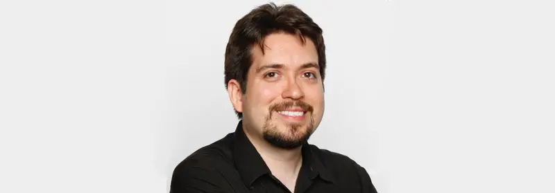 Smiling man with dark hair and a goatee wearing a black shirt against a light gray background