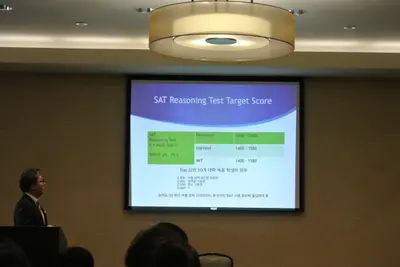 교실에서 화이트보드 앞에 선 교사가 테이블에 앉은 학생들과 이야기를 나누고 있는 모습입니다.