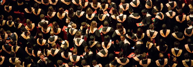 Overhead view of a dense crowd of graduates in black gowns with gold and orange hoods at a commencement ceremony