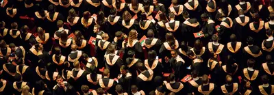 Overhead view of a dense crowd of graduates in black gowns with gold and orange hoods at a commencement ceremony