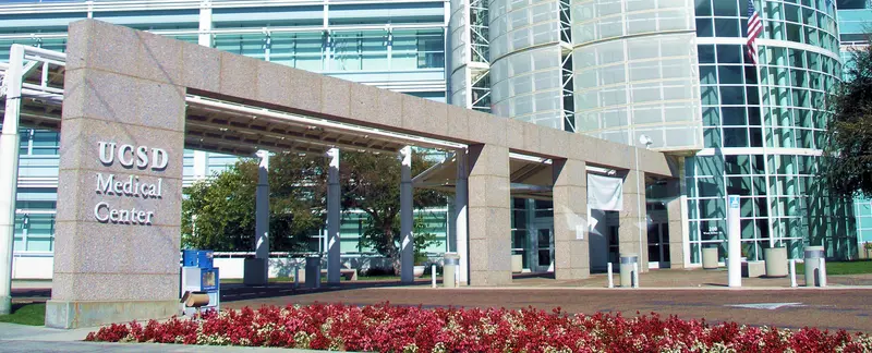 "UCSD Medical Center"라고 적힌 석조 표지판과 유리 건물 입구, 앞쪽에 붉은 꽃이 피어 있는 모습입니다.