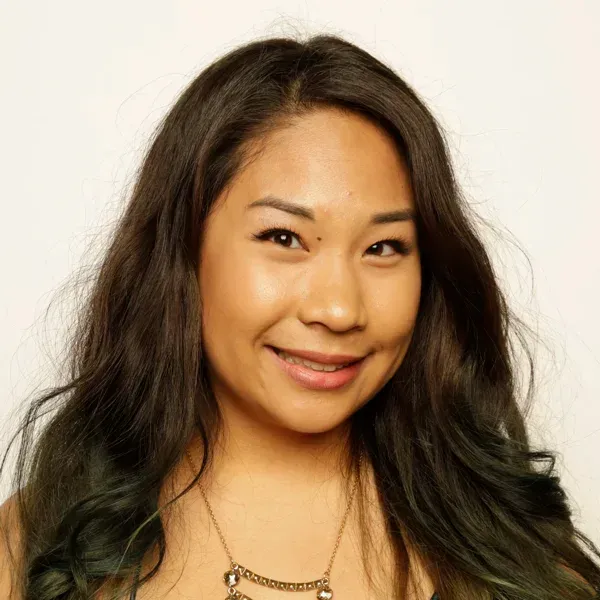 Smiling young woman with long dark wavy hair and a beaded necklace against a plain background