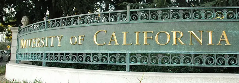 Curved bronze campus sign with gold lettering reading "University of California" amid trees