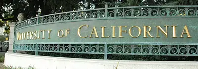 Curved bronze campus sign with gold lettering reading "University of California" amid trees