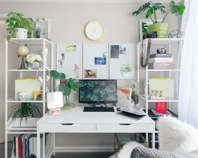 A tidy white home office desk with a monitor, keyboard, plants, shelves and a wall clock above pinned photos.
