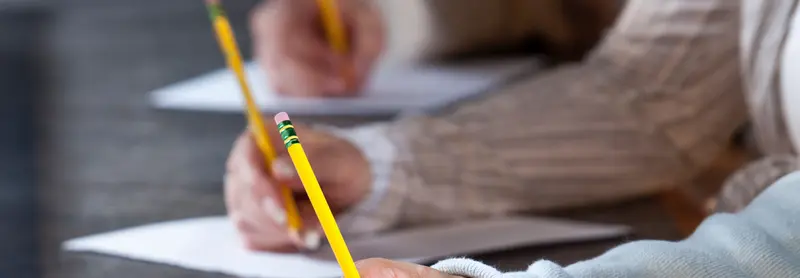 Close-up of two students holding yellow pencils and writing on papers at a desk