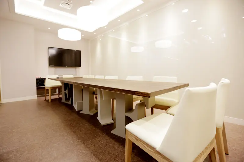 Bright empty classroom with a long wooden table, white chairs, and a wall-mounted flat-screen TV.