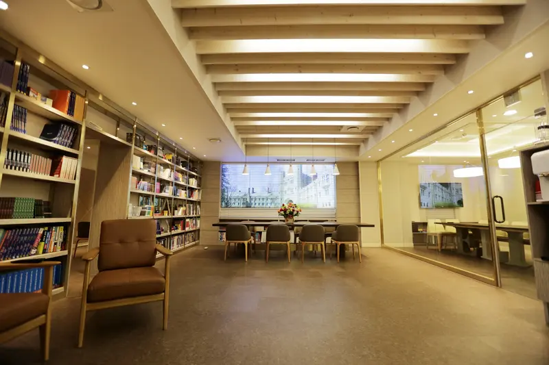 Study center interior with tall bookshelves, armchair, long table and chairs by a window, and exposed wood beam ceiling