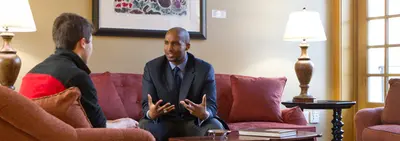 A man in a suit gestures while talking with a student seated across from him in a living room with red sofas.