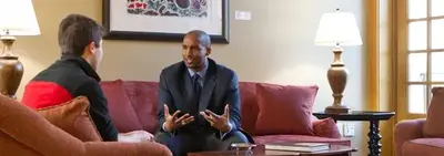 A man in a suit gestures while talking with a student seated across from him in a living room with red sofas.
