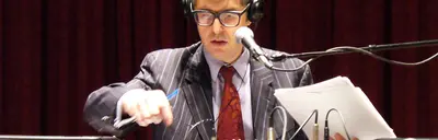 A man in glasses, headphones and a pinstriped suit speaks into a microphone while holding papers and a pen.