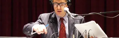 A man in glasses, headphones and a pinstriped suit speaks into a microphone while holding papers and a pen.