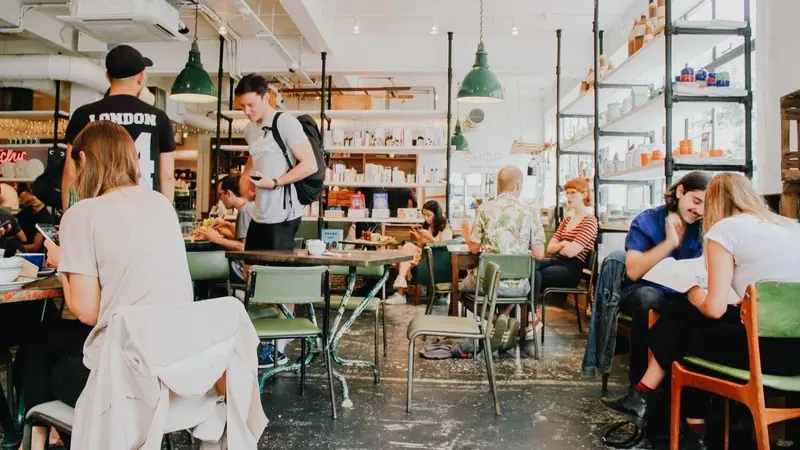 A busy cafe with people seated at tables talking, eating and working, with green pendant lamps overhead.