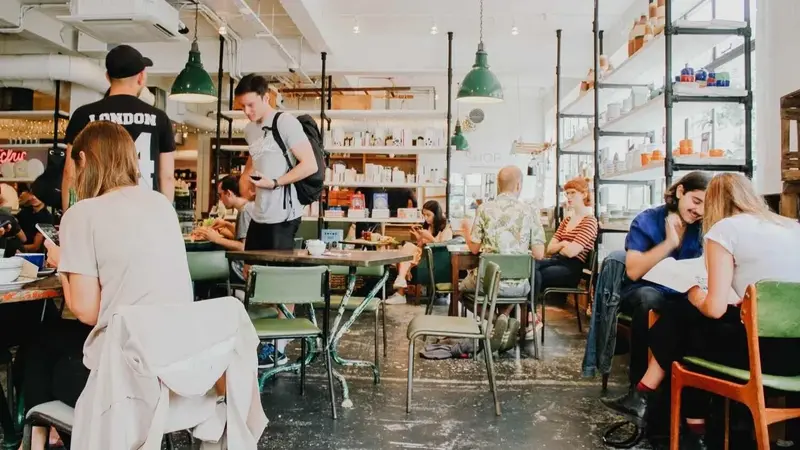 A busy cafe with people seated at tables talking, eating and working, with green pendant lamps overhead.