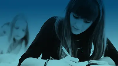 A student writing in a notebook, a classmate studying behind her