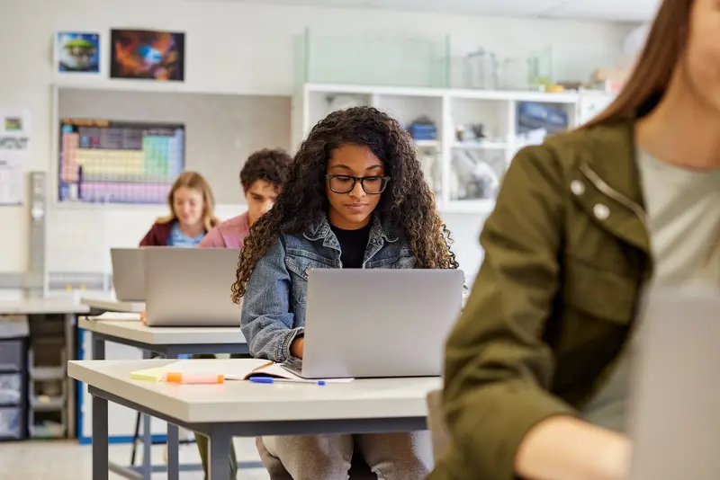 Students working on laptops at desks in a classroom, with a periodic table poster on the wall