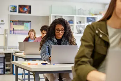Students working on laptops at desks in a classroom, with a periodic table poster on the wall