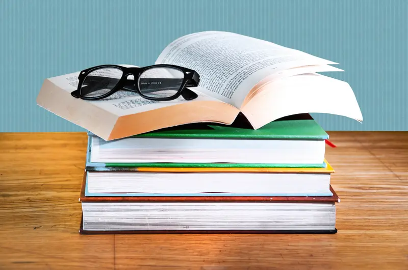 An open book with black-framed glasses resting on it, atop a stack of textbooks on a wooden table