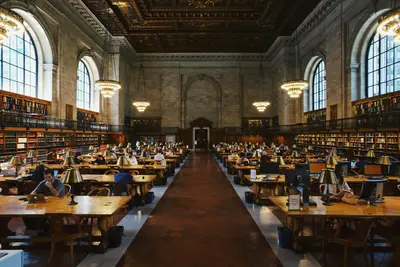 Students studying at long wooden tables with brass lamps in a grand library reading room with chandeliers and arched windows.