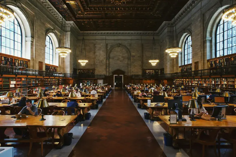 Students studying at long wooden tables with brass lamps in a grand library reading room with chandeliers and arched windows.