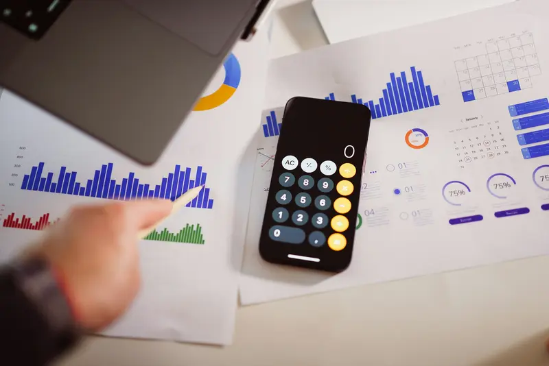 A hand with a pen points at printed bar charts and calendars beside a smartphone showing the calculator app.