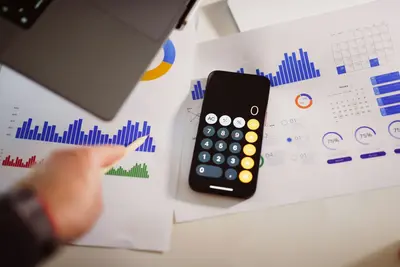 A hand with a pen points at printed bar charts and calendars beside a smartphone showing the calculator app.