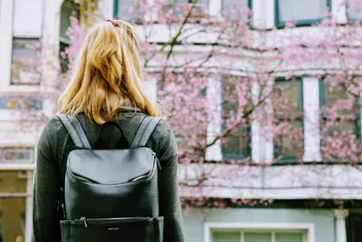 A blonde student seen from behind wearing a black leather backpack, facing a building and blossoming pink tree