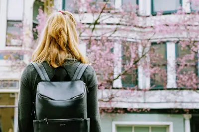 A blonde student seen from behind wearing a black leather backpack, facing a building and blossoming pink tree