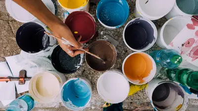 Overhead view of a hand stirring a bucket of brown paint surrounded by open buckets of colorful paint