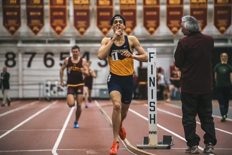 A runner in a Concordia singlet sprints past a FINISH marker on an indoor track as another athlete trails behind.