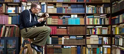 A man in glasses sits on a step ladder reading a book in front of floor-to-ceiling shelves of books.