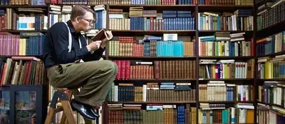 A man in glasses sits on a step ladder reading a book in front of floor-to-ceiling shelves of books.